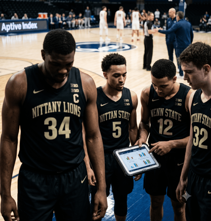 Four basketball players in dark uniforms gather on the court; one player wearing a captain's "C" patch stands in front while three teammates look at a digital tablet showing charts and data.